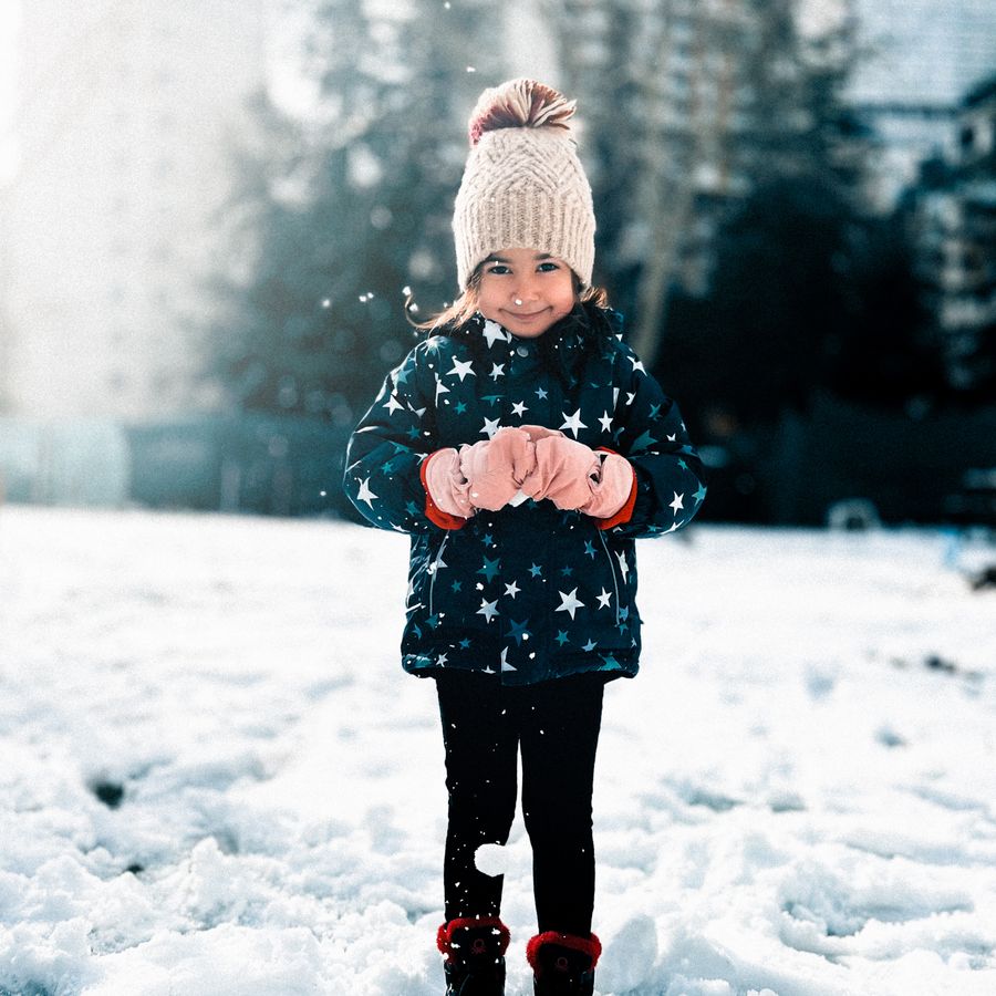 A child standing in fresh snow in a winter coat, knitted hat and mittens.